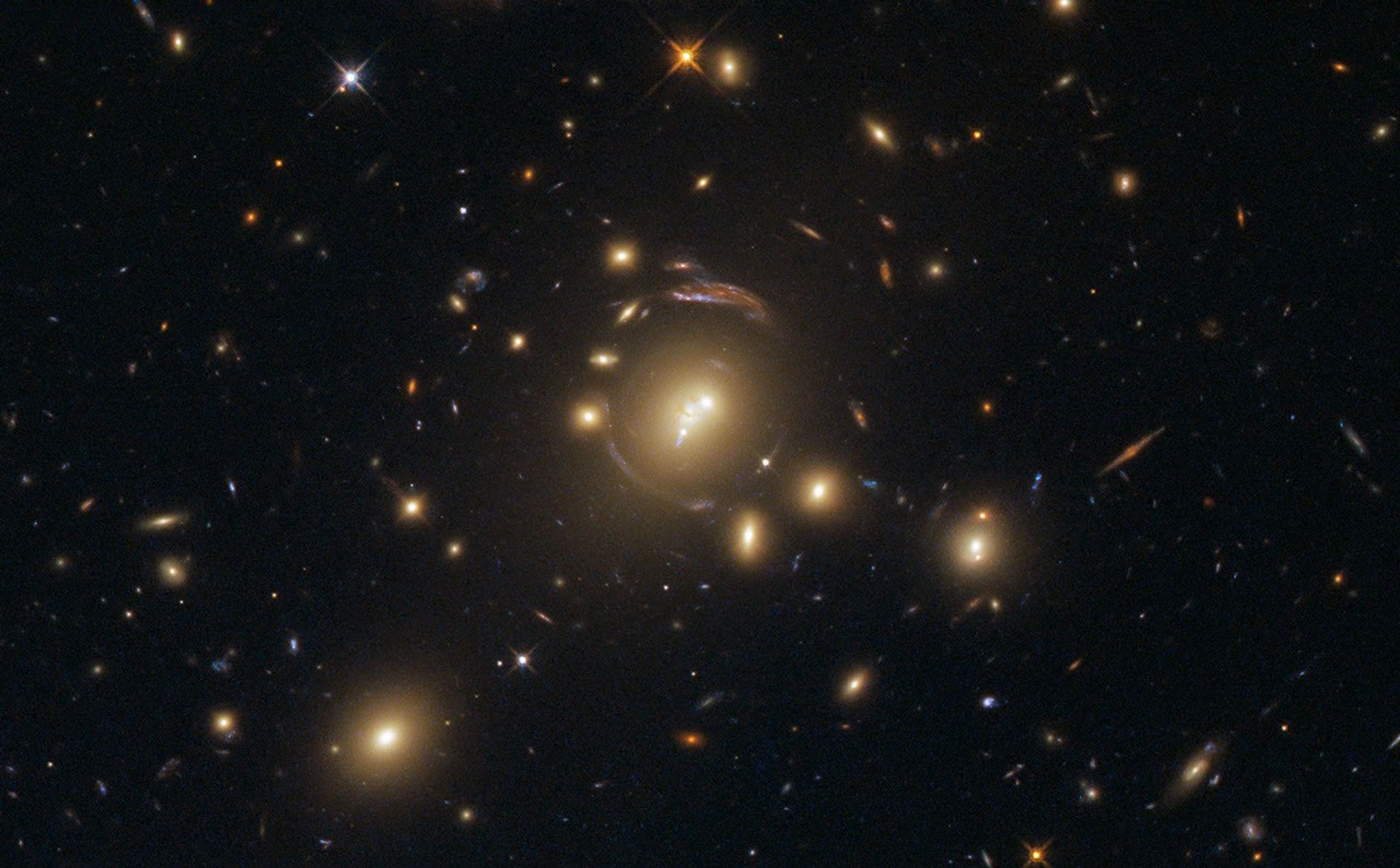 Black background dotted with galaxies. A reddish group of galaxies at image center with a brownish-red ring curving almost completely around it.